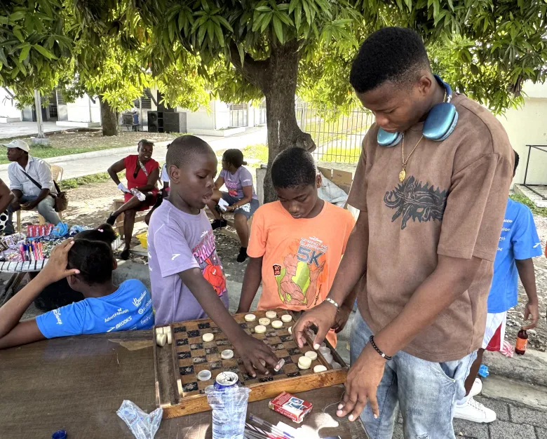 A young man and a child play checkers during “Let’s Play Together” on Saturday, Aug. 15, 2026, at Lycée Anacaona in Léogâne, Haiti. Photo by Daniella Saint-Louis for The Haitian Times. 
