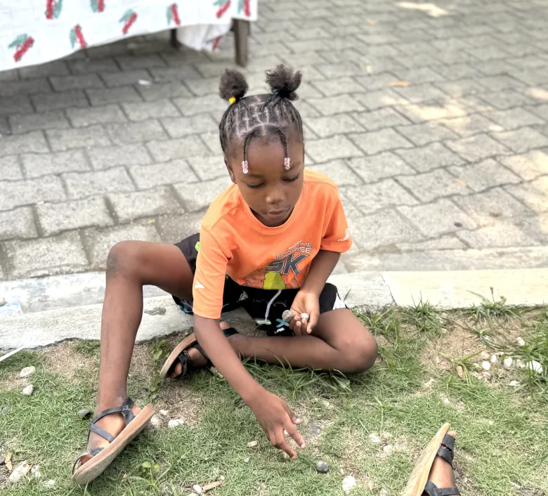 Une petite fille pratique le jeu d’osselets avec de petits cailloux lors de “Let’s Play Together” le samedi 15 août 2026, au Lycée Anacaona à Léogâne, Haïti. Photo de Daniella Saint-Louis pour The Haitian Times. 

